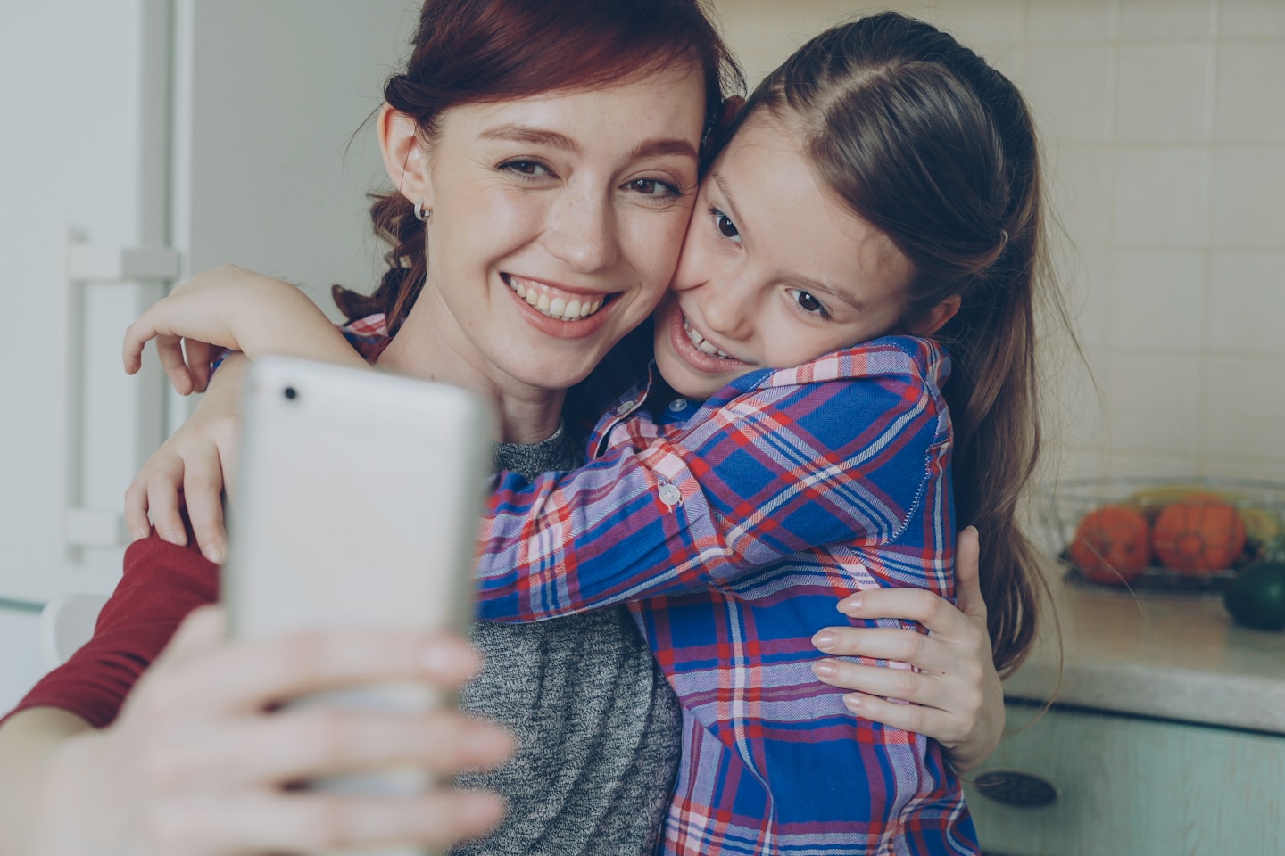 Smiling mother together with funny playful daughter making selfie photo with smartphone camera at home in kitchen. Family, cooking, and people concept