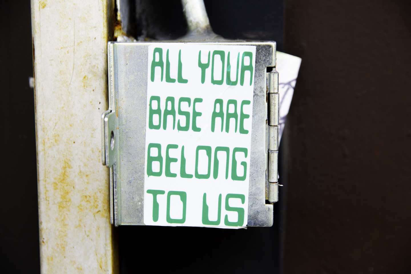 A clean white sticker attached to a metal utility box reads: “ALL YOUR BASE ARE BELONG TO US.” The phrase references the famous internet meme and early gaming culture, transforming an overlooked city surface into a piece of nostalgic public humor.