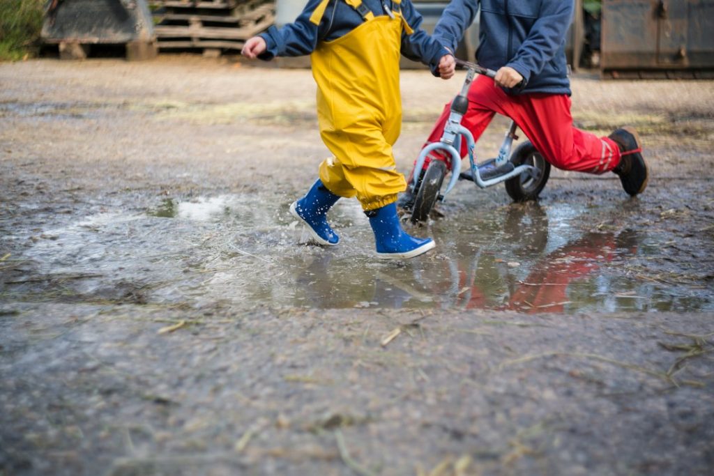 Deux enfants en pantalon de pluie et bottes jouant dans une flaque avec une draisienne